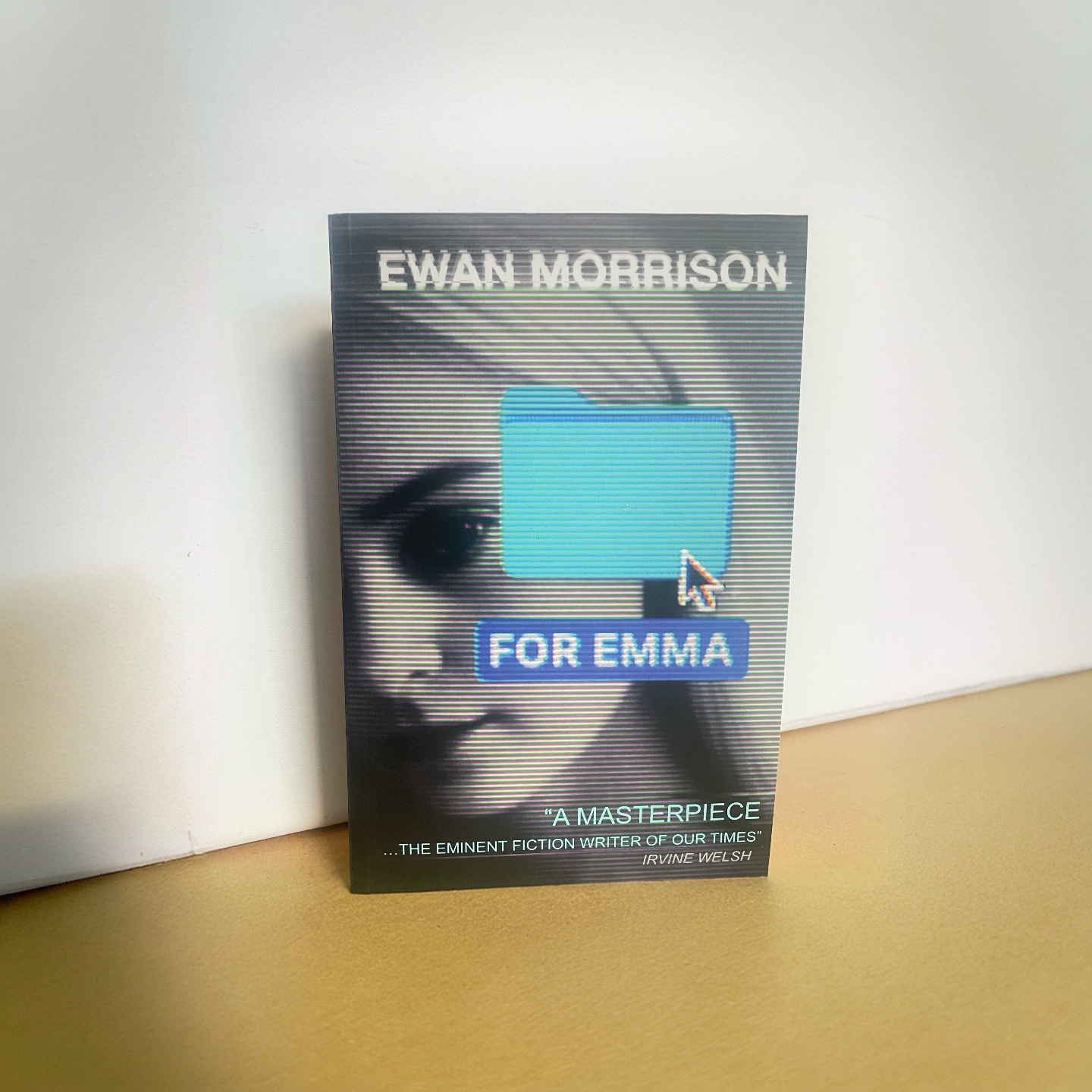 the cover of the novel For Emma by Ewan Morrison shows a blue desktop folder with a mouse pointer hovering over it, the folder is named FOR EMMA and the desktop background is a young woman's face, she has white hair and is white skinned with a dark eye, the words 'a masterpiece... the eminent fiction writer of our times, quoted from Irvine Welsh are printed at the foot, and the author's name, Ewan Morrison, is written at the top in a glitched out light blue font