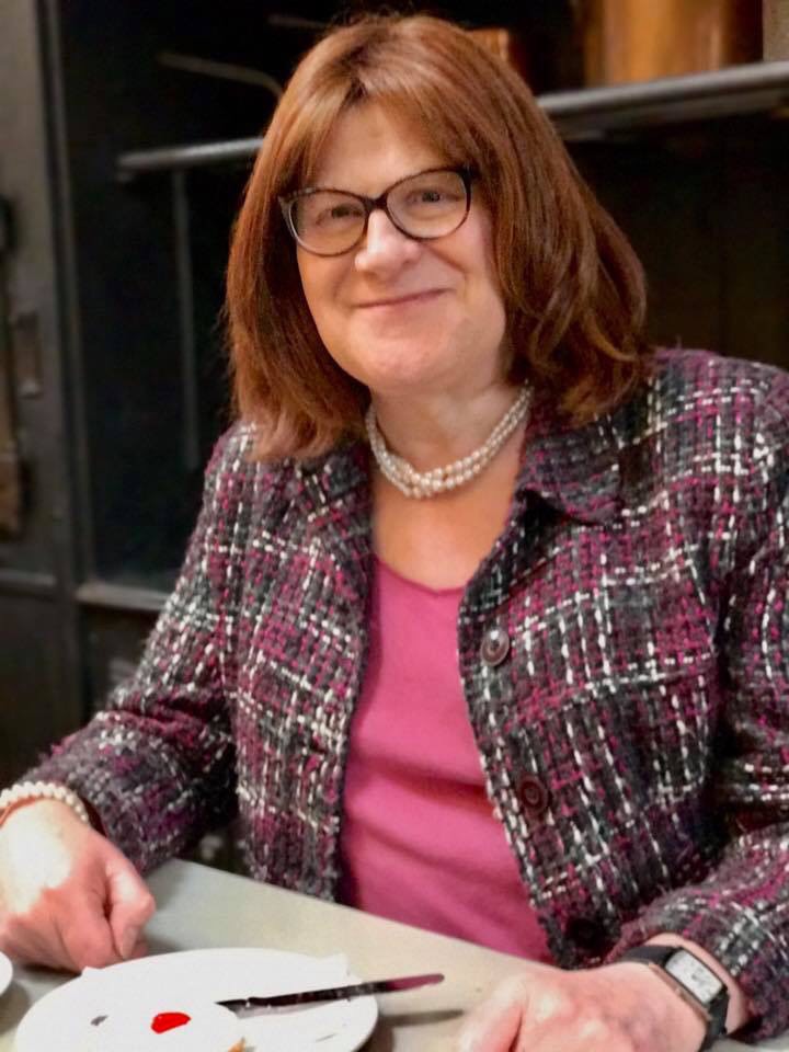 Writer Lesley Storm smiles, she sits a table, with a single Bakewell tart before her, she wears a pearl necklace and a magenta top, and purple coat.