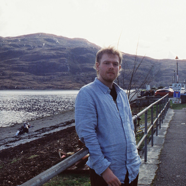 author jo higgs in a light blue shirt leans against a railing in front of the shore of a Scottish loch